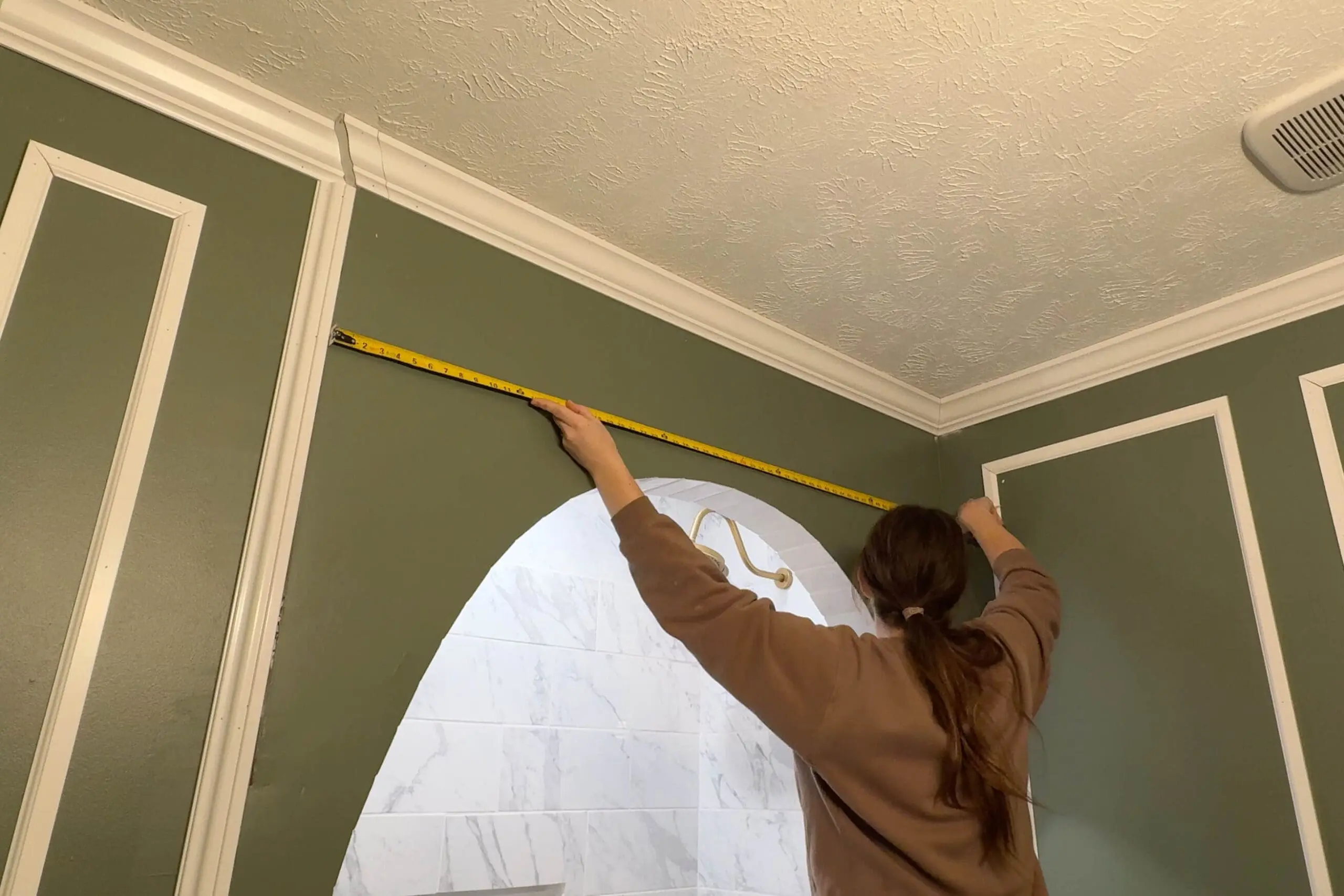 DIY bathroom renovation in progress with a person measuring a green wall for precise trim placement. The archway leads to a tiled shower area with white marble-like tiles and a luxurious gold showerhead.